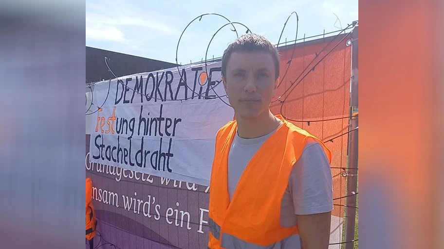 Simon Schwan vor dem Banner samt Stacheldraht am Zaun des Demokratiefests.