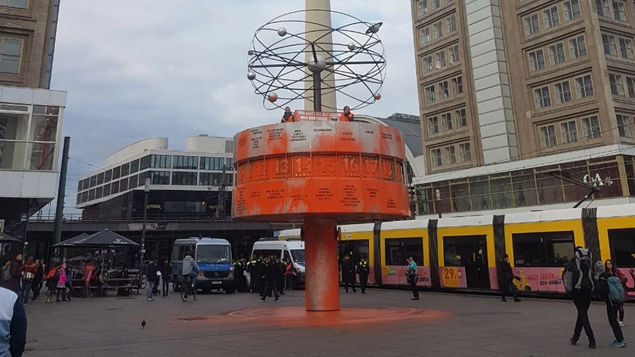 Unterstützer:innen der Letzten Generation haben die orange gefärbte Weltzeituhr bestiegen. Dort entrollten sie ein Banner mit der Aufschrift 